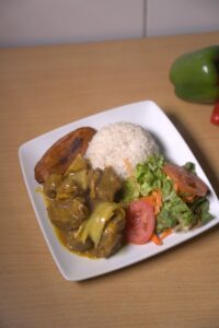 A plate of Jamaican curried goat served with white rice, fried plantain, and fresh salad with lettuce, carrots, and tomatoes.